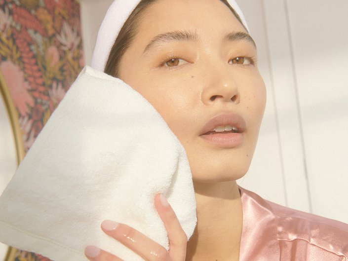 Person wearing a white headband and pink satin robe blotting their cheek with a white washcloth